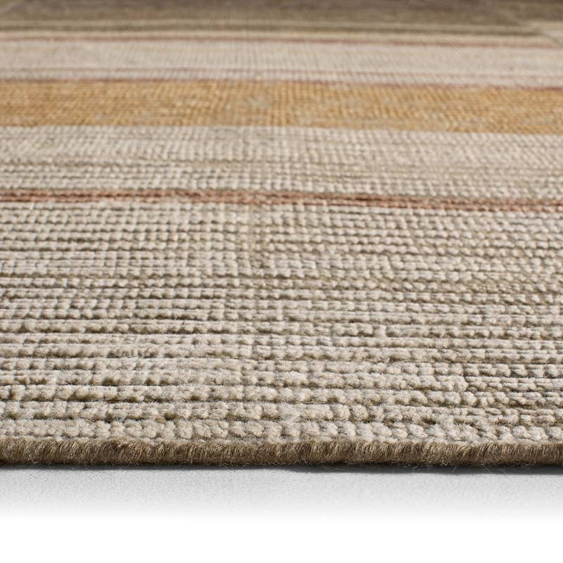 A close-up of a woven area rug with horizontal stripes in beige, cream, tan, and light brown, showing the detailed texture of the fibers and a fringed edge.