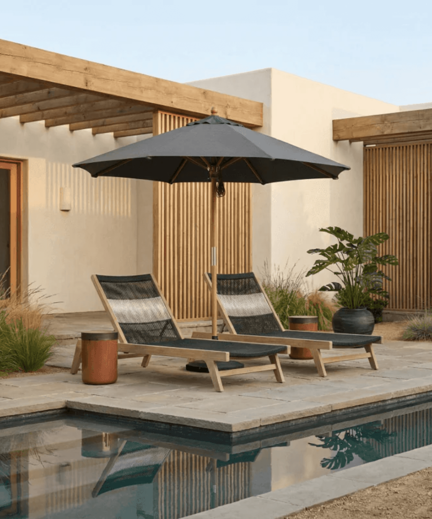 Two lounge chairs under a large umbrella by a poolside with a modern building in the background.