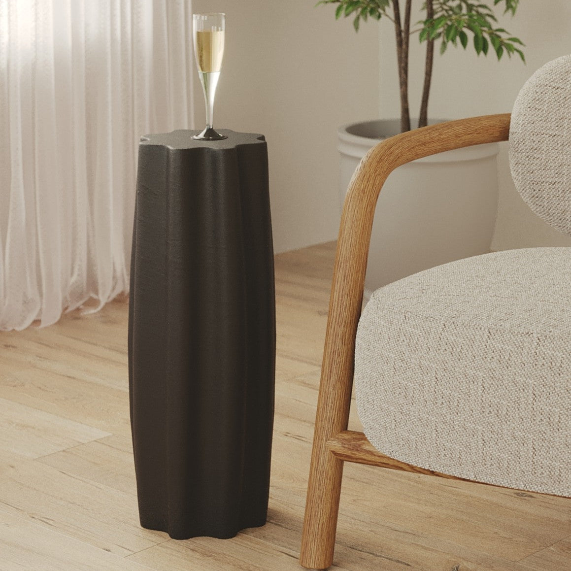 A dark charcoal gray fluted pedestal side table with a single stem glass on top, styled in a bright living room next to a light wood armchair.