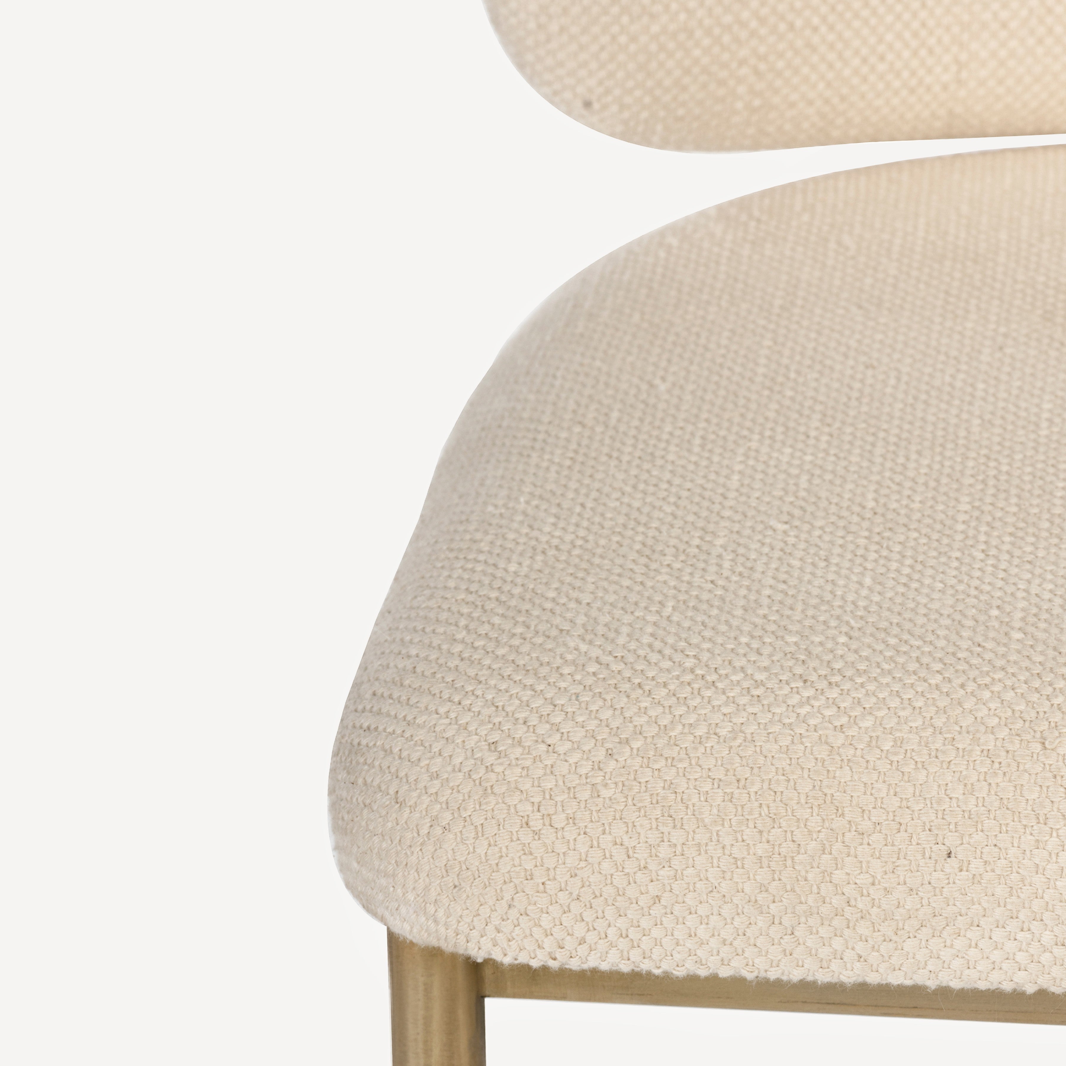 Close-up of a woven beige upholstered chair seat with a small portion of the matching curved backrest and a brass metal leg visible against a white background.