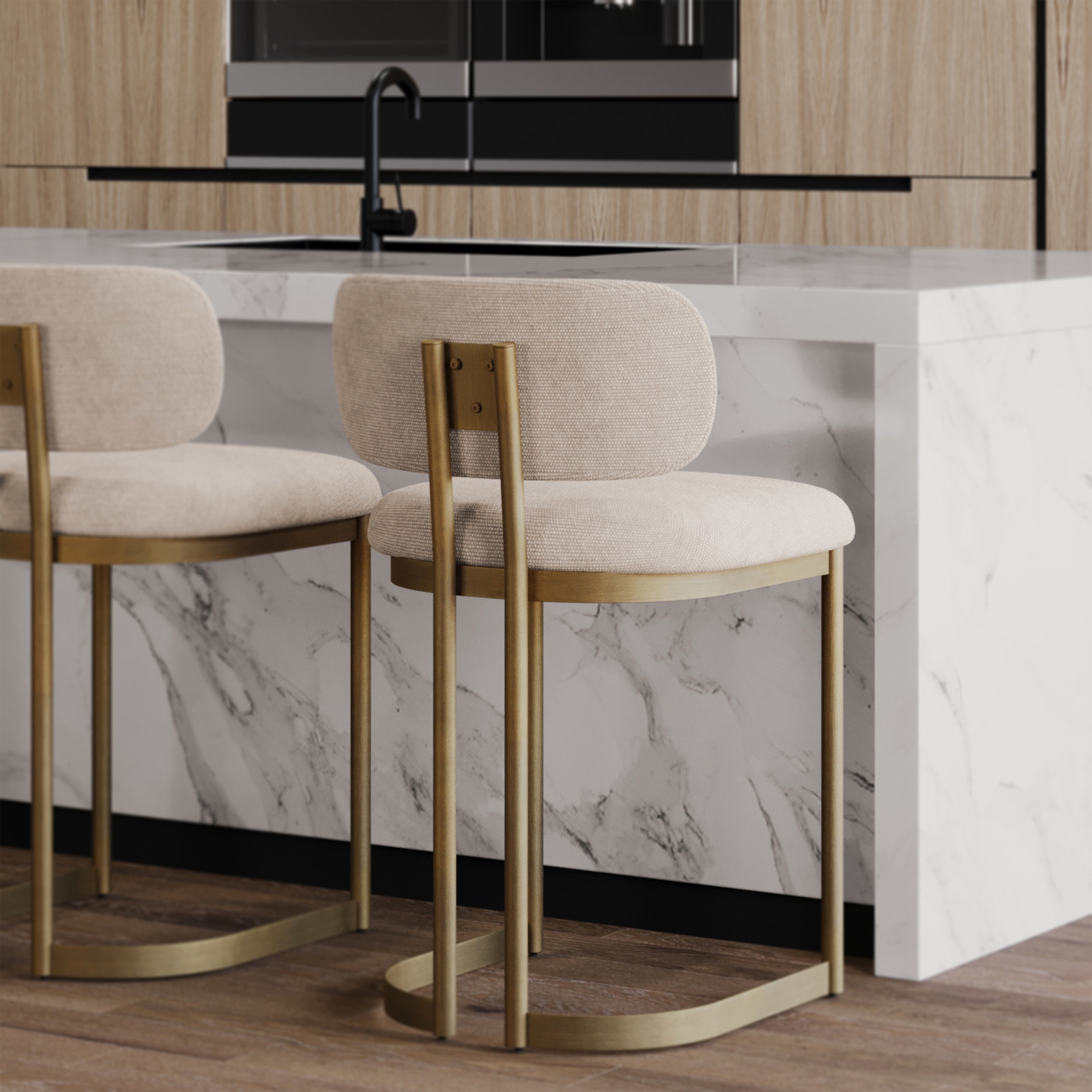 Two modern bar stools with beige upholstered seats and backs, slim gold metal frames, and curved gold footrests, positioned at a white marble kitchen island in a contemporary kitchen setting.