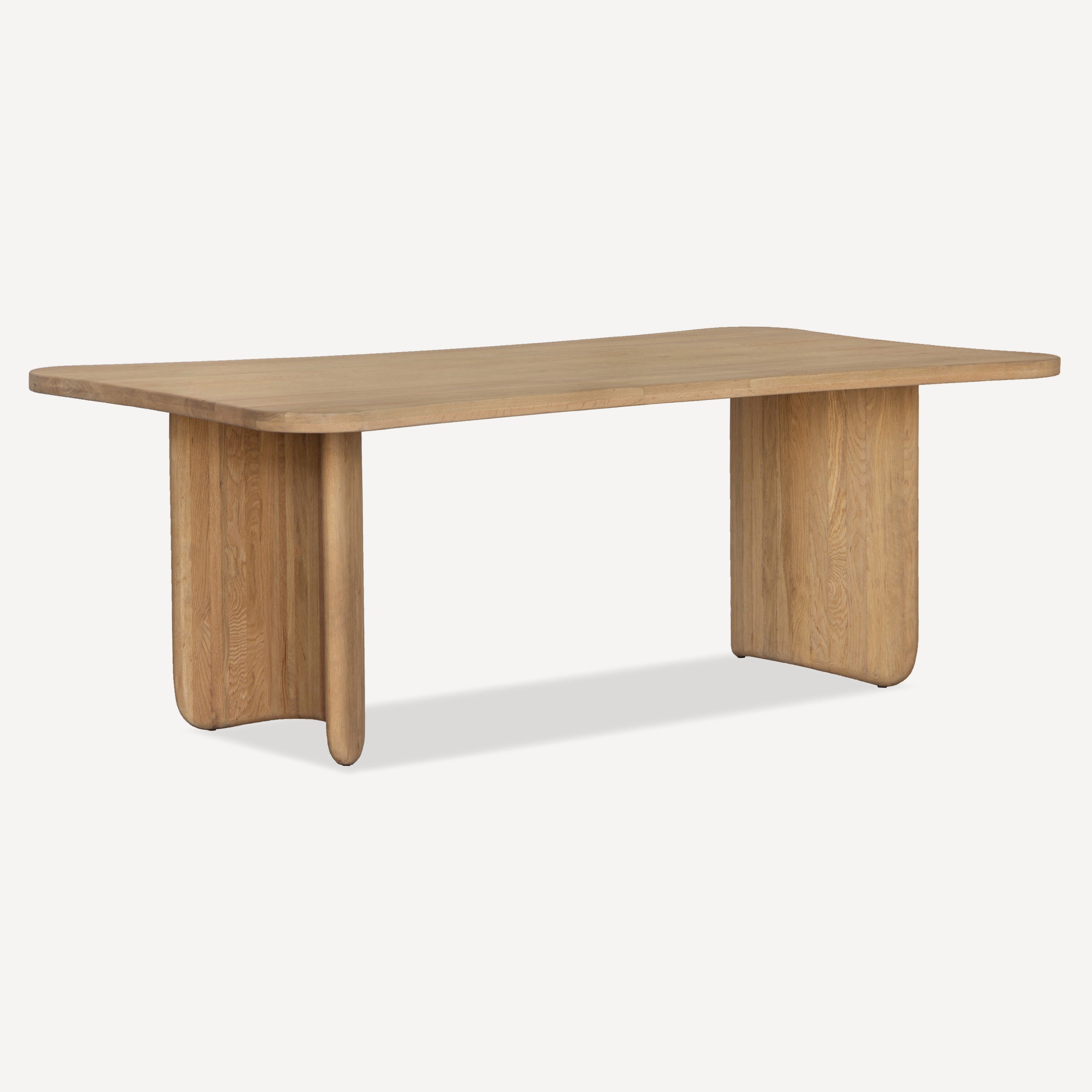 A modern rectangular wooden table with a natural oak finish, featuring thick block-style legs and rounded corners, shown on a light gray background.