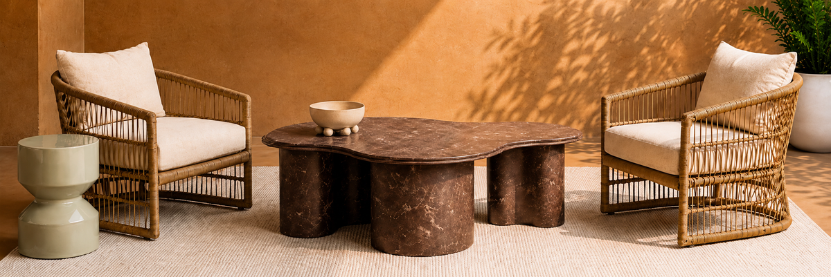A styled outdoor patio furniture set featuring two woven rattan armchairs with cream cushions, a matching round coffee table with a dark brown stone top, and a small ceramic bowl, set against a warm terracotta wall with leaf shadows.