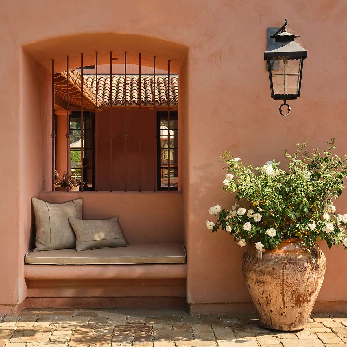 Outdoor seating area with a cushioned bench against a pink wall, featuring a potted plant and wall-mounted lantern.