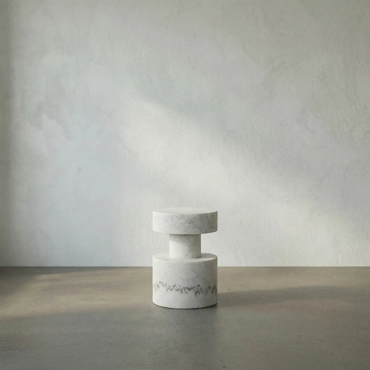 A minimalist two-tiered white stone stool with a textured finish and a small dark floral accent near the base, set against a neutral gray concrete floor and textured wall.