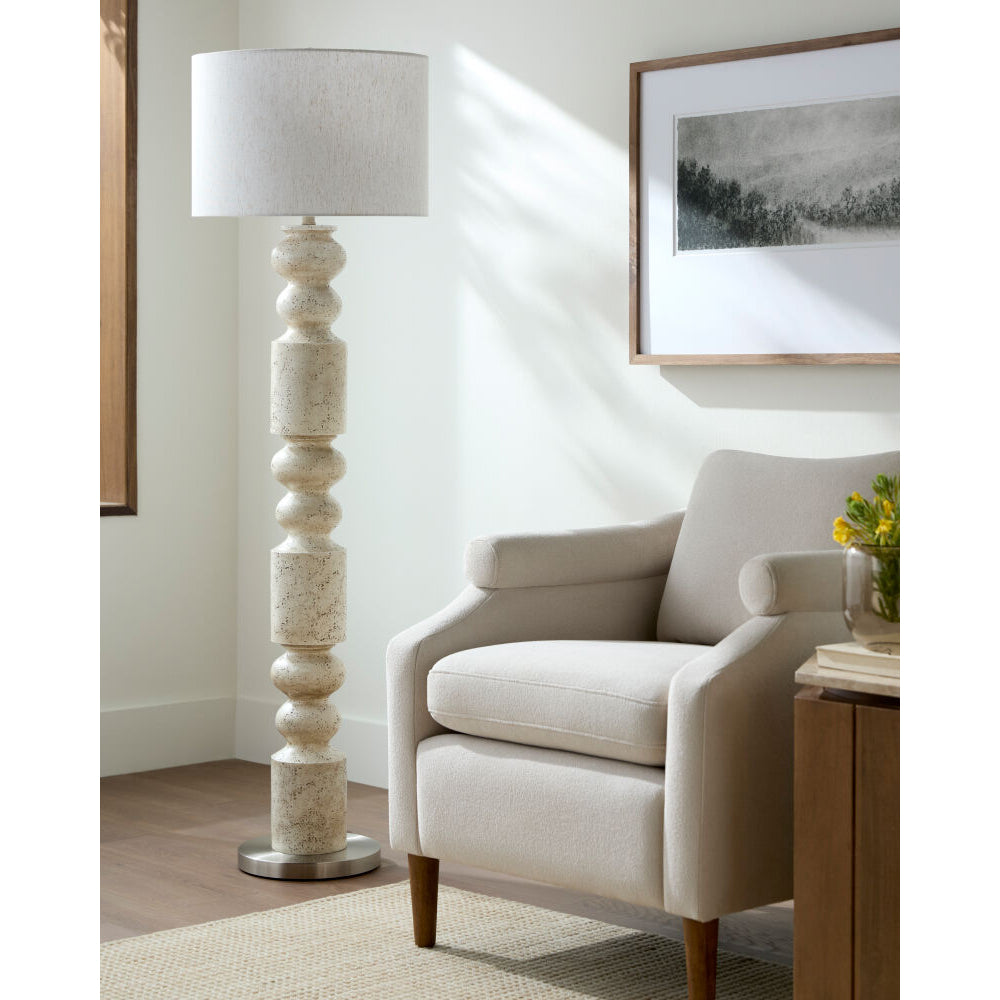 Beige armchair in a living room with a tall lamp and framed picture on the wall.