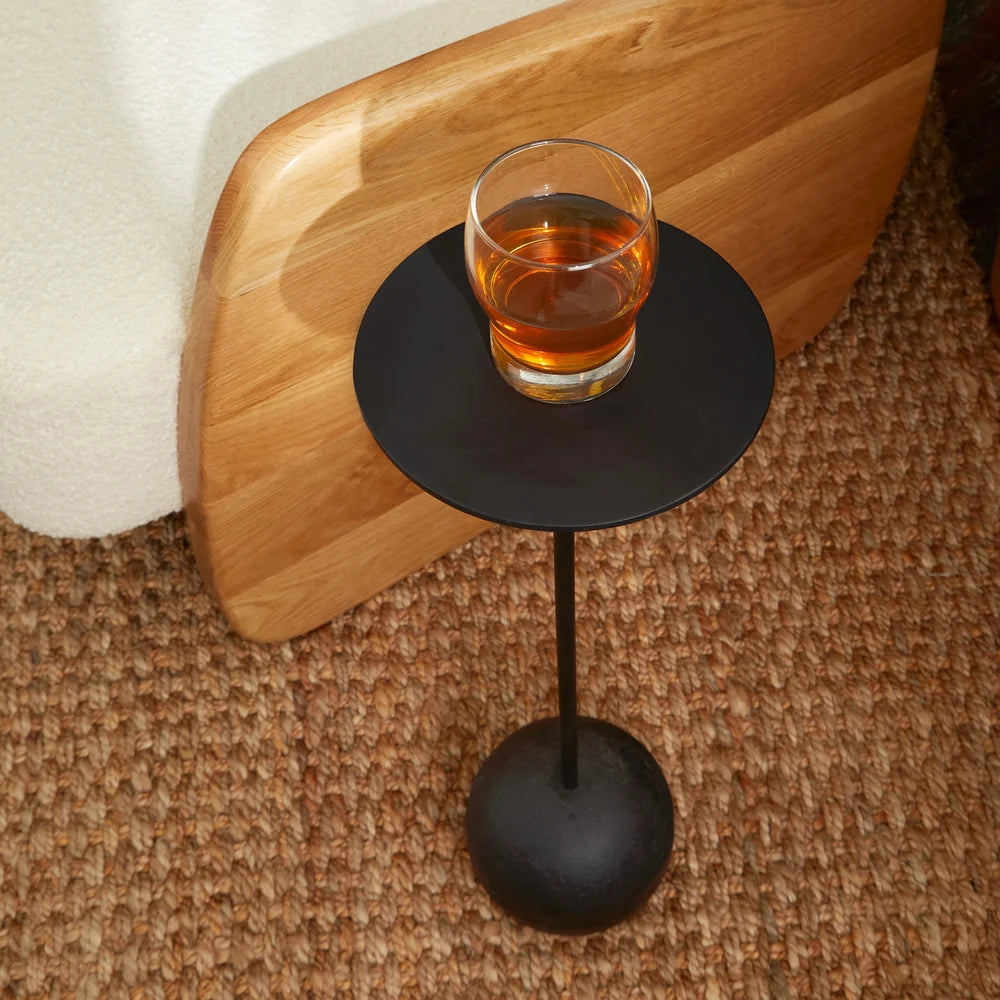 A small round black metal side table with a single pedestal base and a clear glass of amber liquid on top, placed on a woven brown rug beside a wooden board and white textured surface.