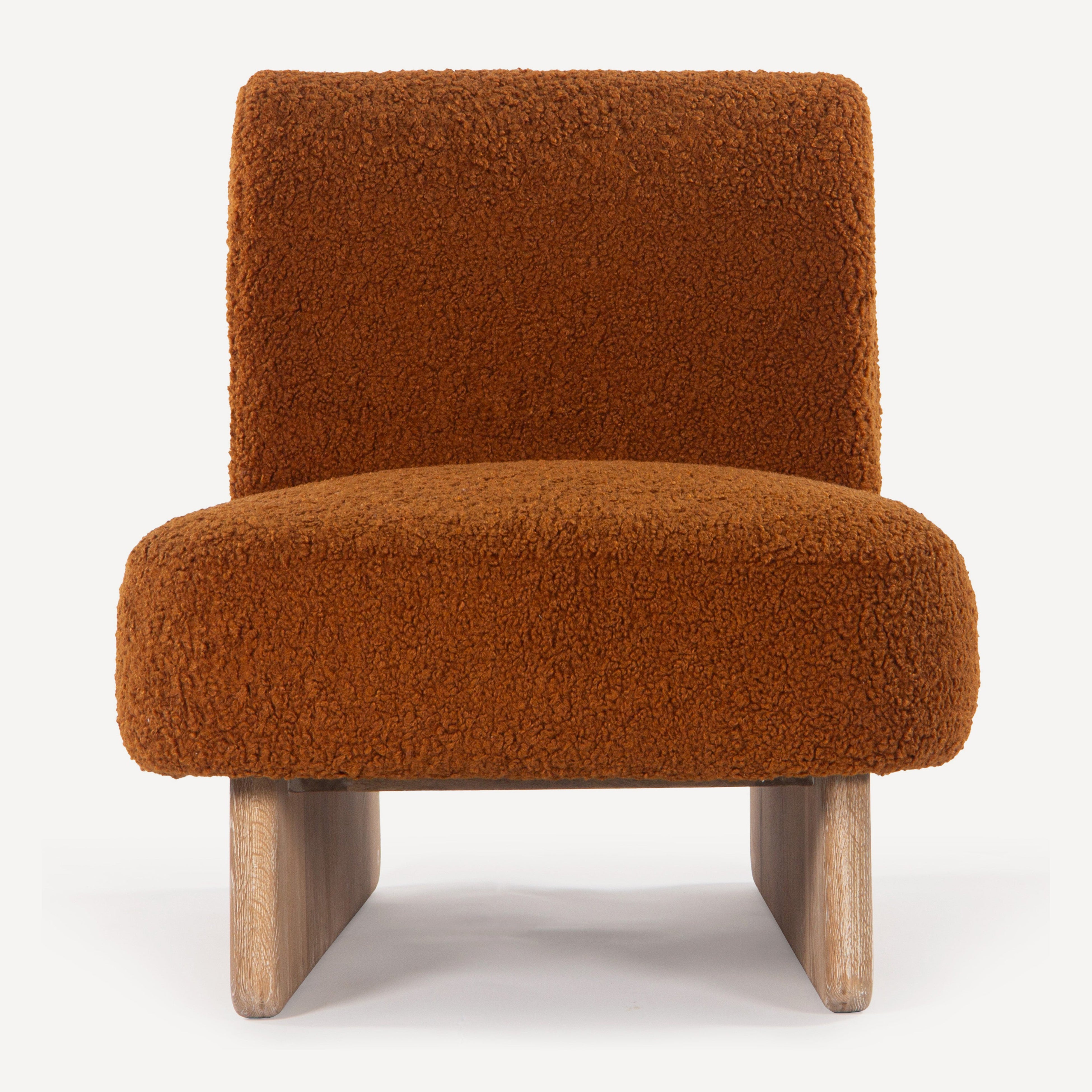 A modern low-profile armless accent chair upholstered in a warm rust-brown bouclé fabric, supported by light natural wood legs, shown against a plain white background.