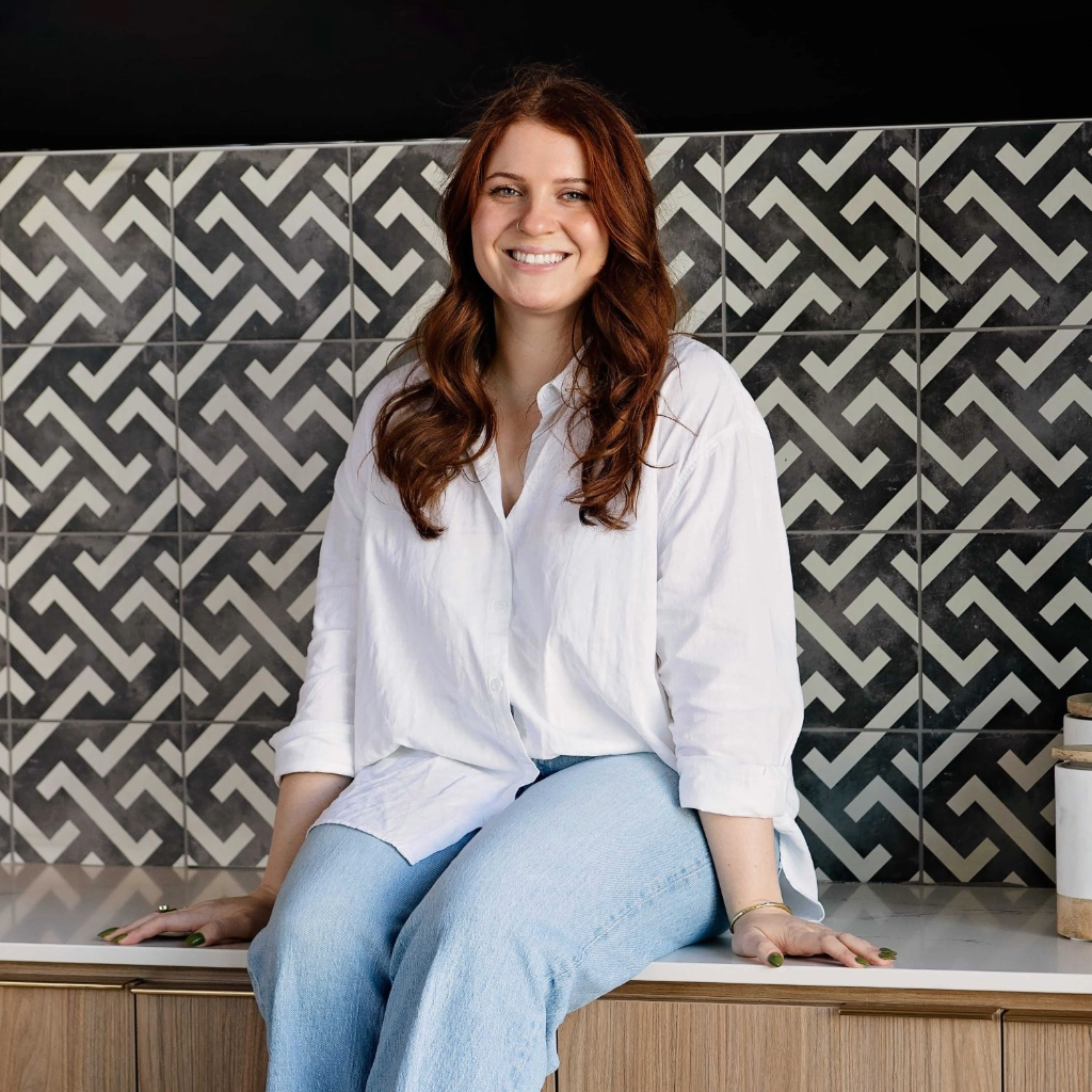 A smiling woman with long wavy brown hair sits on a kitchen counter, wearing a white button-up shirt and light blue jeans. The background features a black and white geometric patterned tile backsplash.