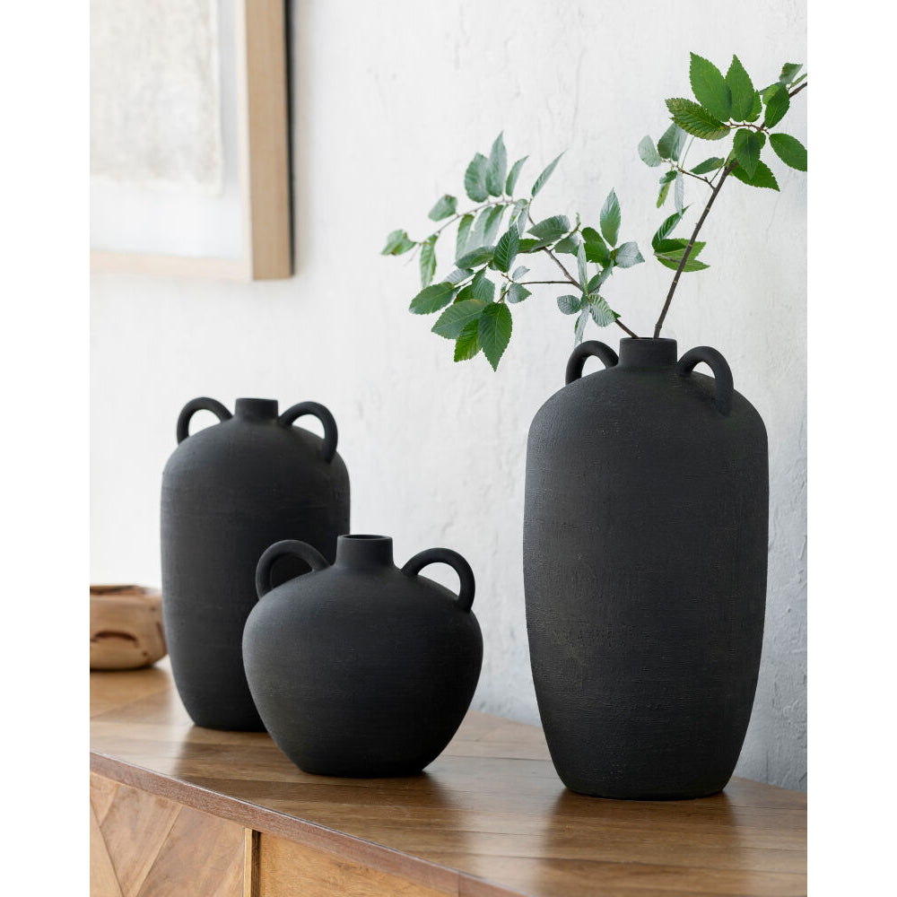Three black vases on a wooden surface with a plant in the background