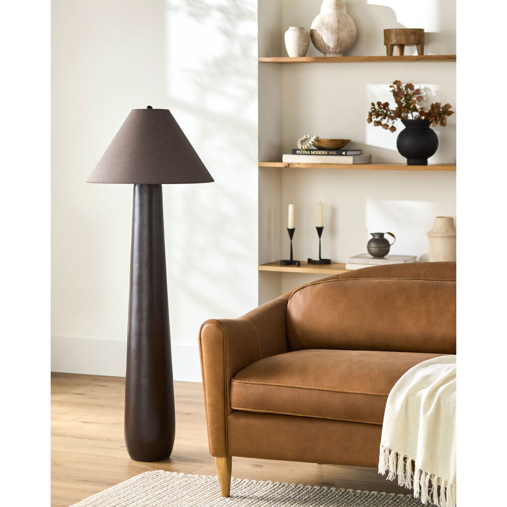 A modern floor lamp with a tapered dark brown shade and a sleek, tapered dark brown base stands next to a brown leather sofa in a styled living room setting.