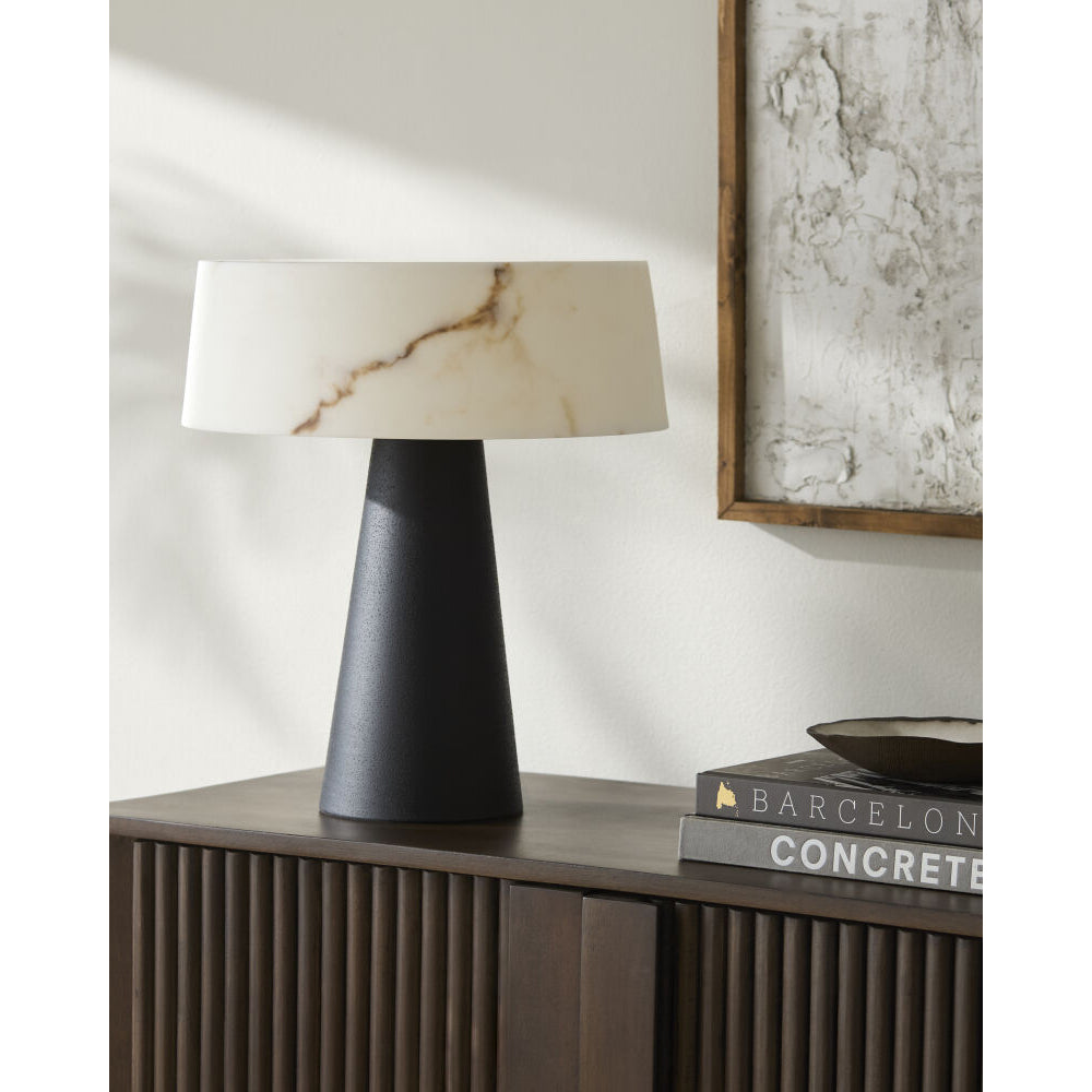 A modern table lamp with a matte black tapered base and a white stone drum shade featuring subtle warm brown veining, styled on a dark wood console table next to books.