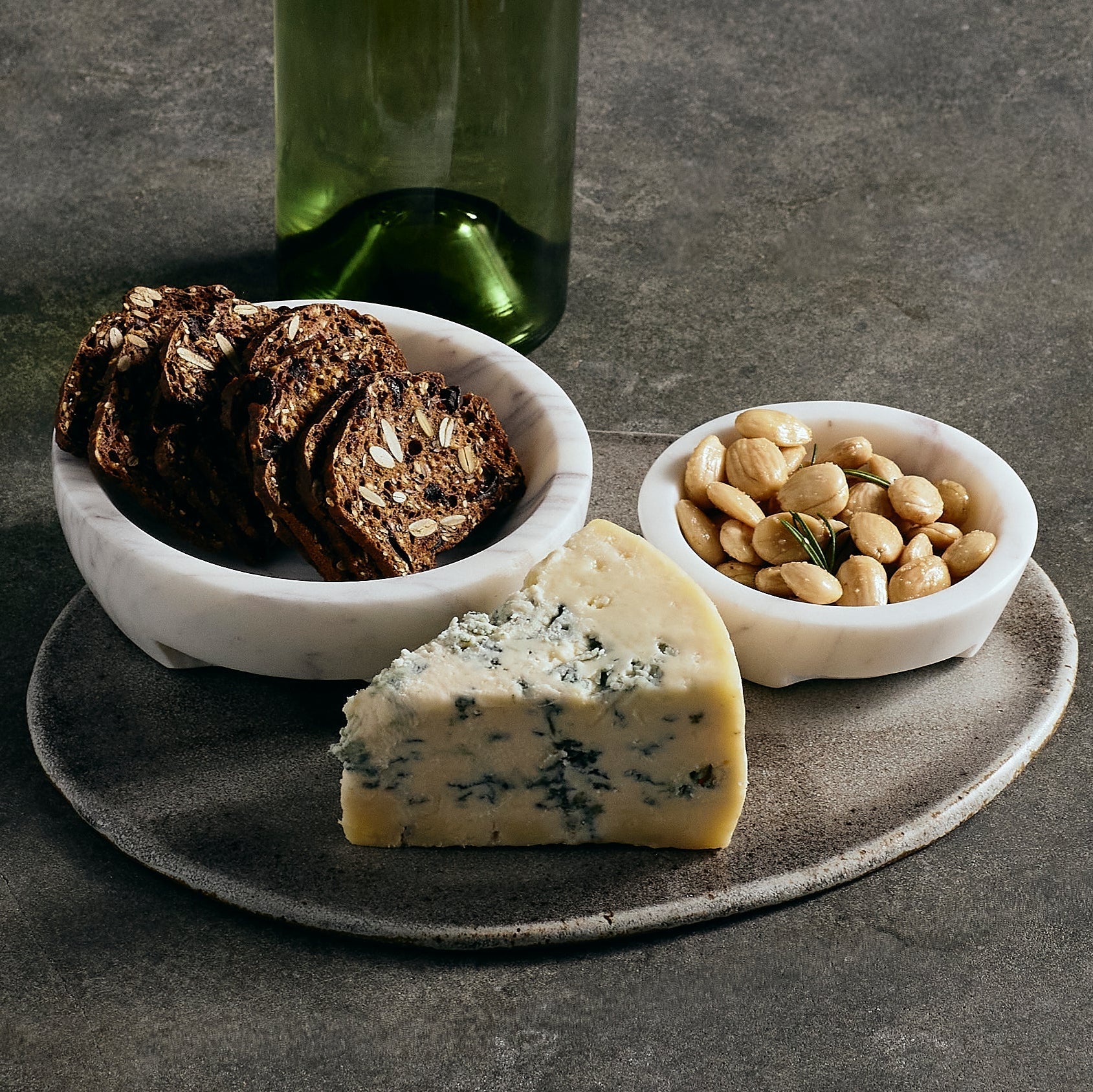 Platter with marble bowls filled with blue cheese, crackers, and nuts on a dark surface