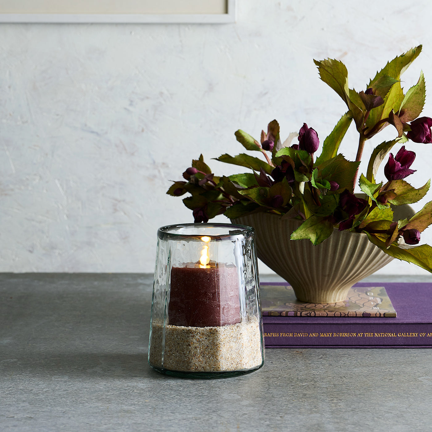 Candle in a glass holder with sand and flowers in a vase on a table.