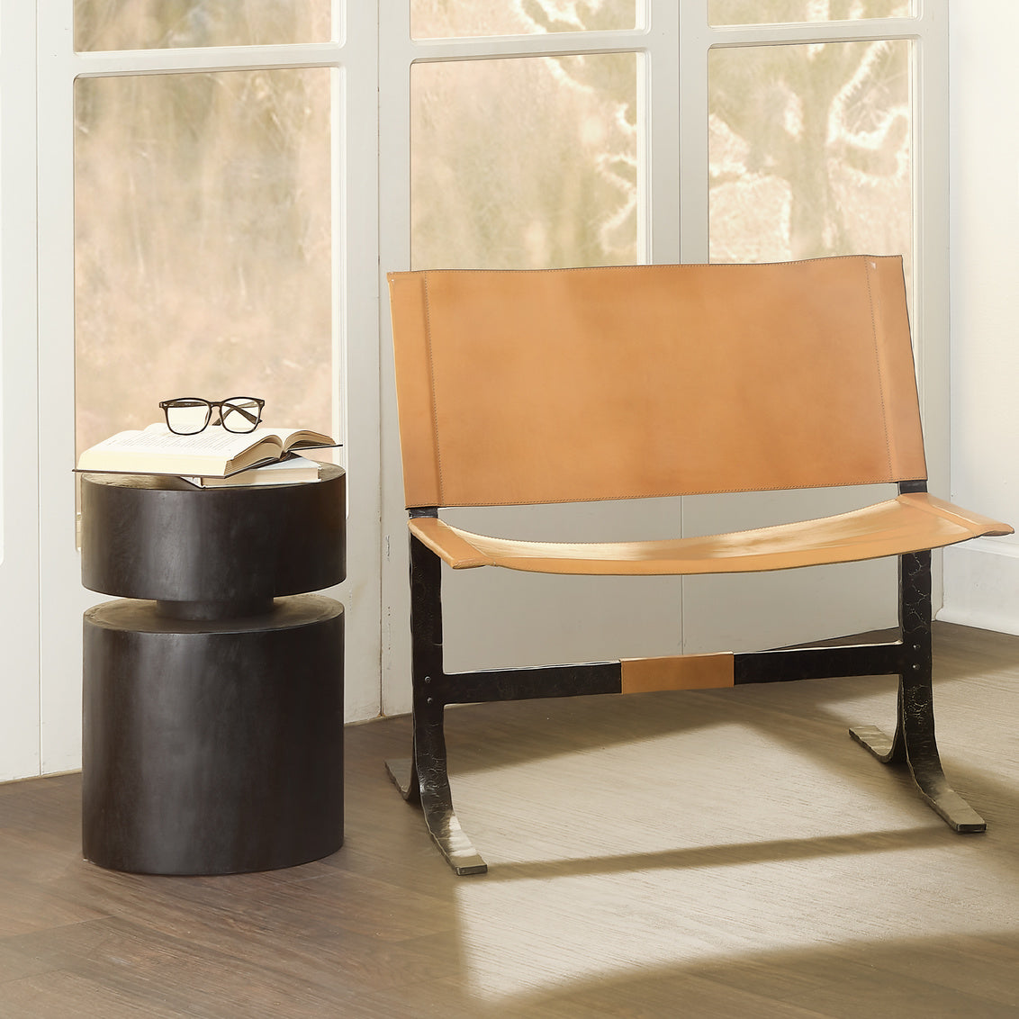 Brown leather bench and black cylindrical side table with glasses and book in a room with large windows.