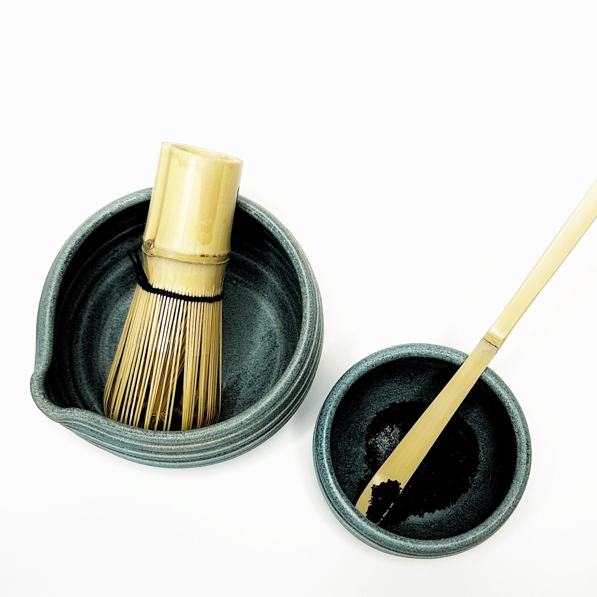 Two small black ceramic bowls with bamboo whisk and spoon on a white background