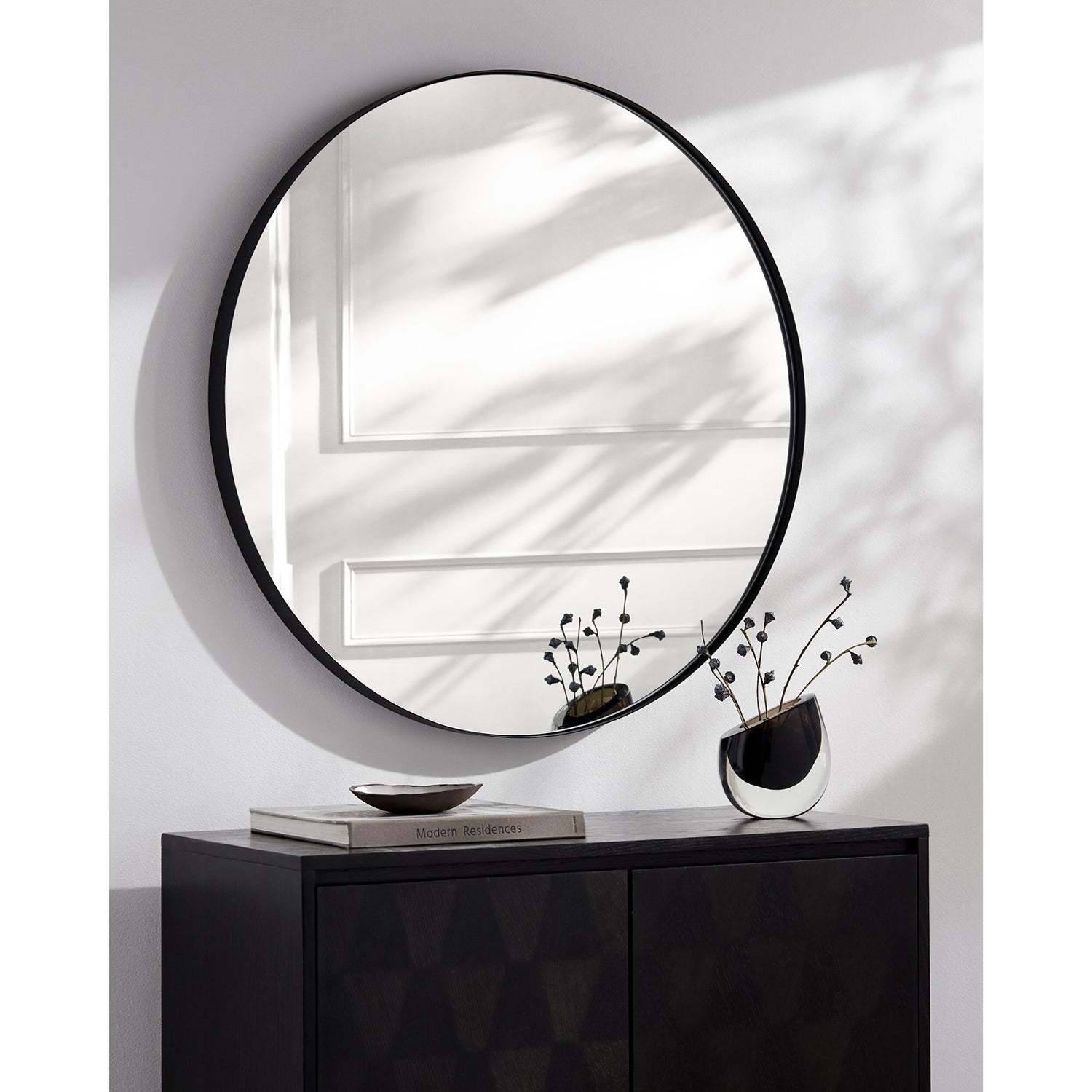 Round mirror with black frame on a white wall above a dark cabinet with decorative items.
