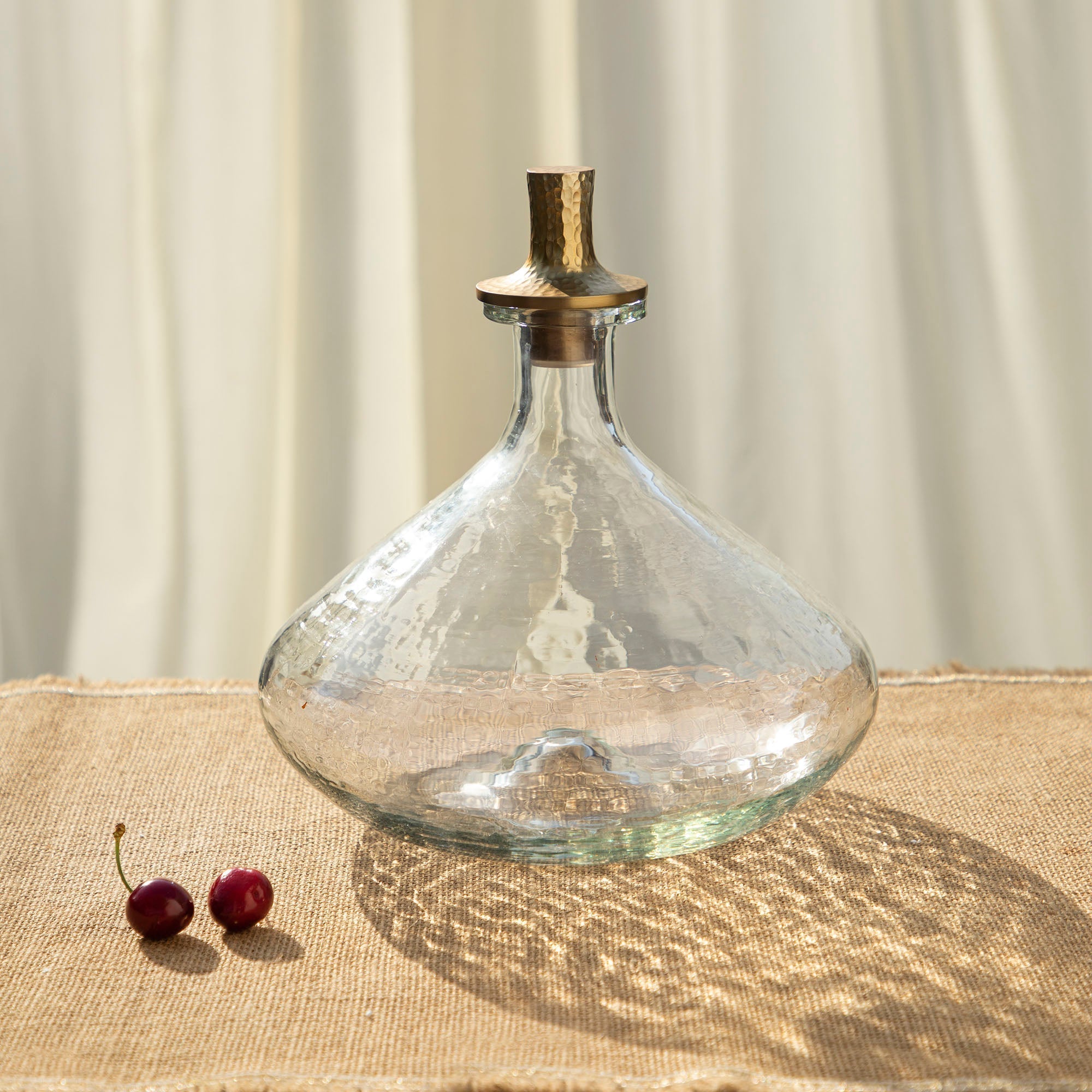 Clear glass decanter with a gold stopper on a textured surface with two cherries.