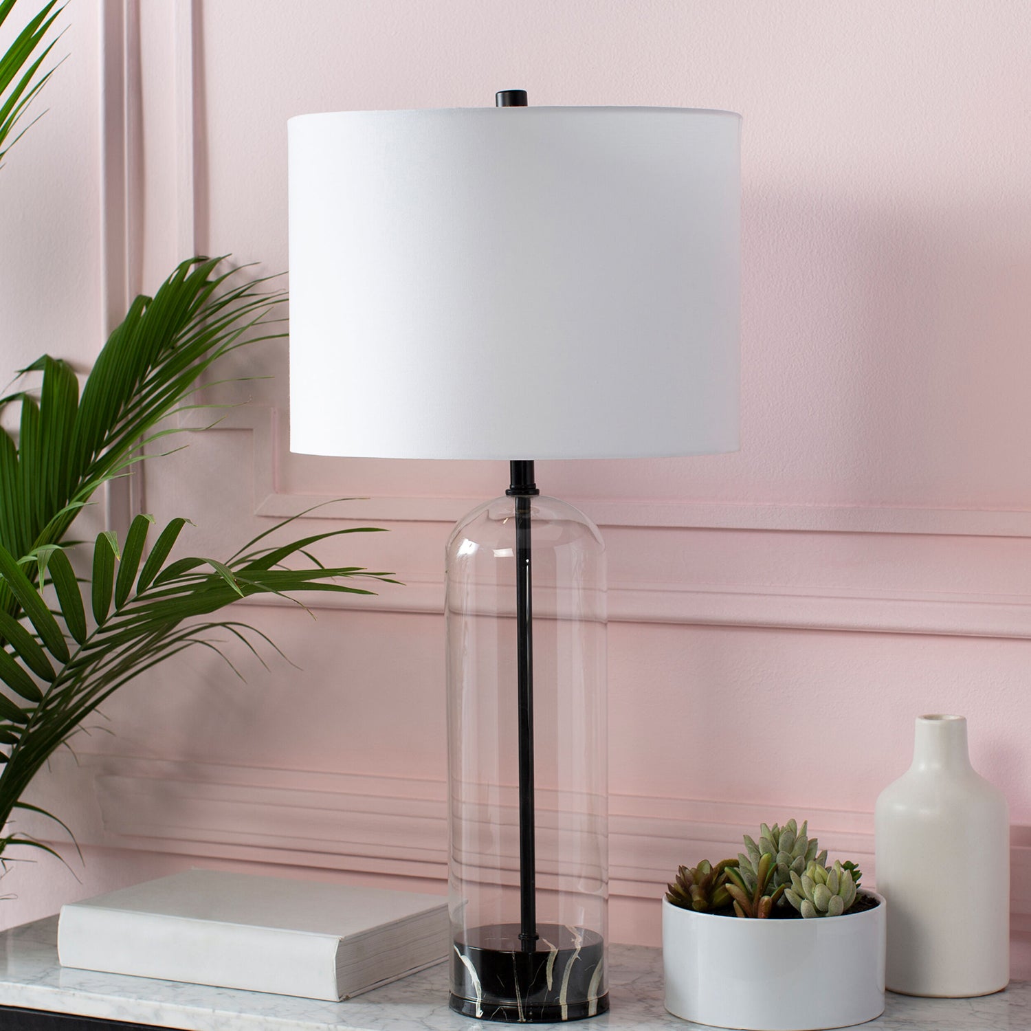 Table lamp with white shade and clear cylindrical base on a surface with books and plants against a pink wall.