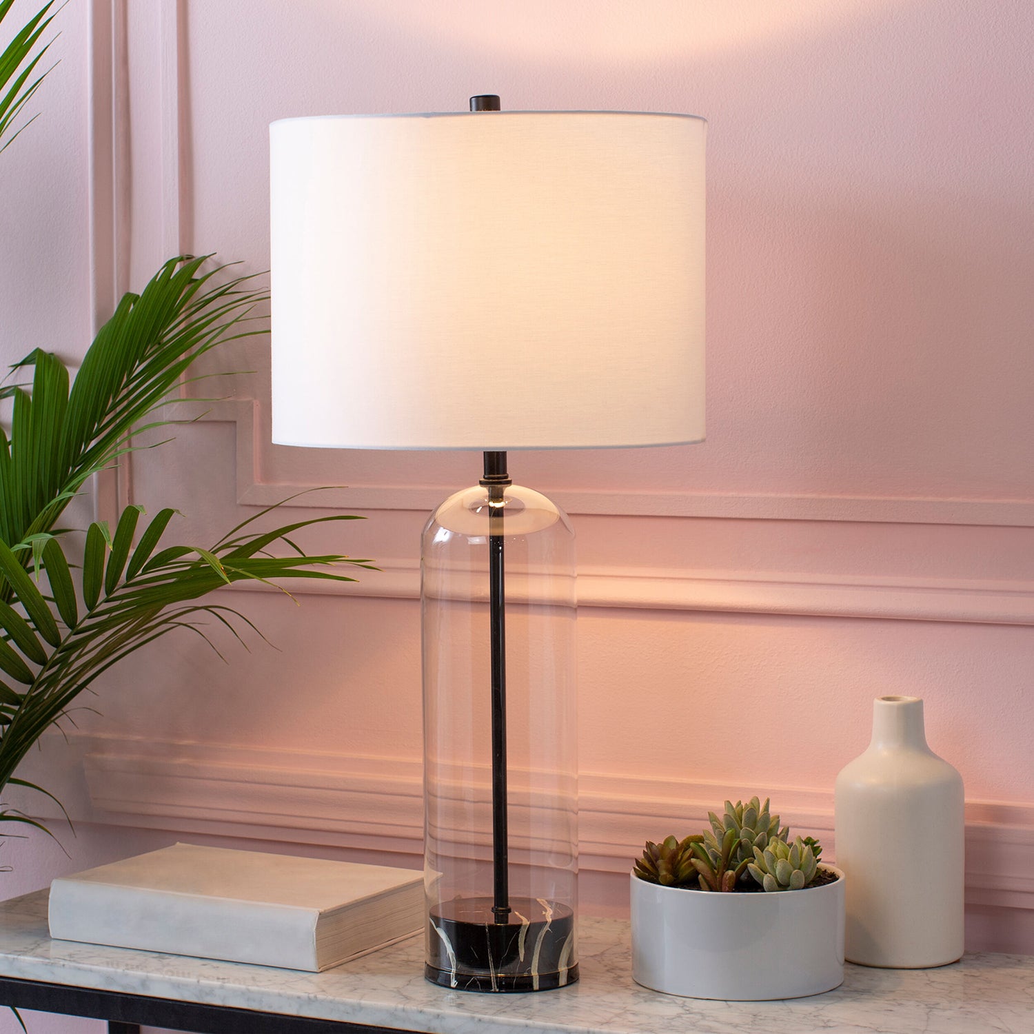 Table lamp with a white shade on a marble surface next to a plant and small vase.