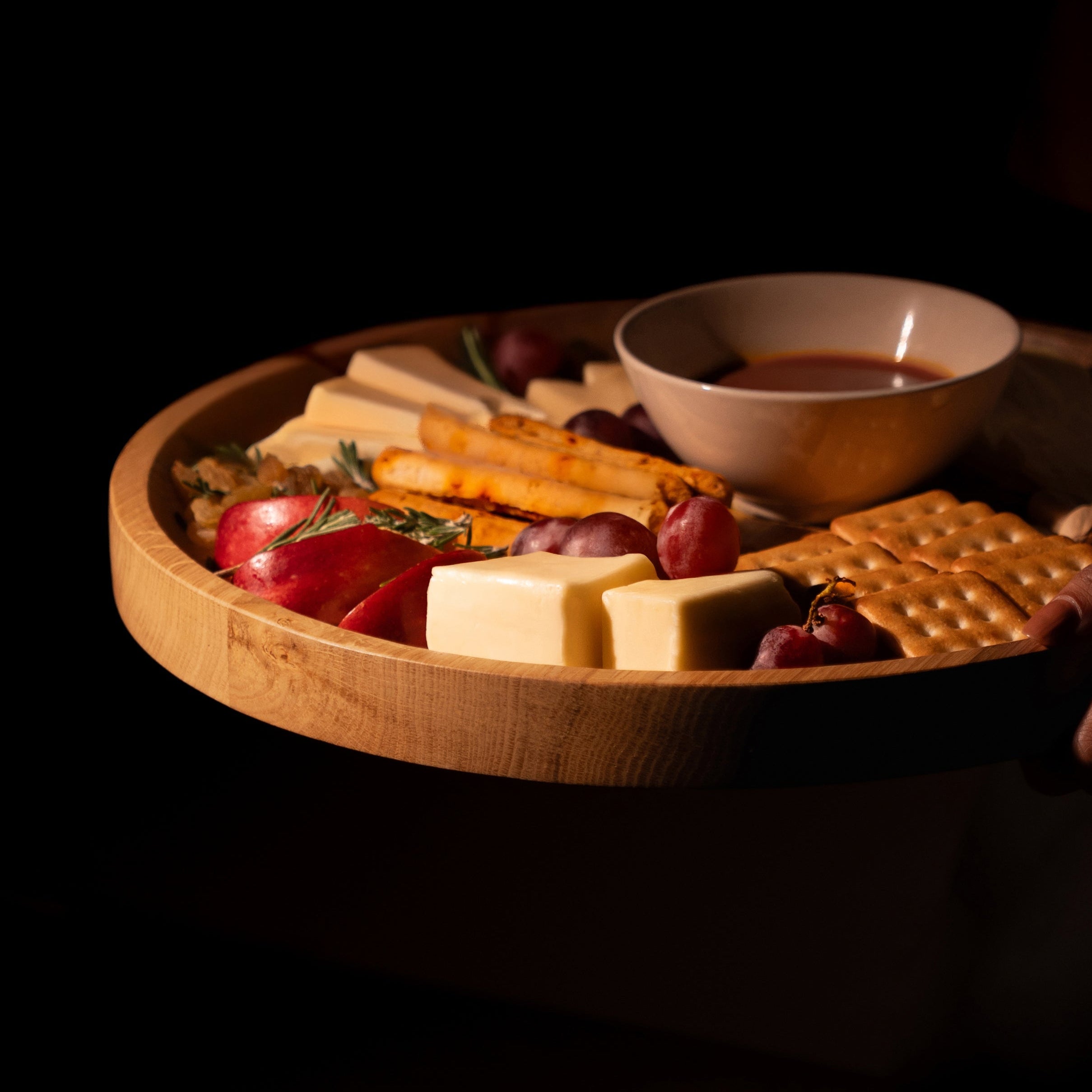 Wooden tray with assorted snacks including crackers, cheese, and fruits on a dark background