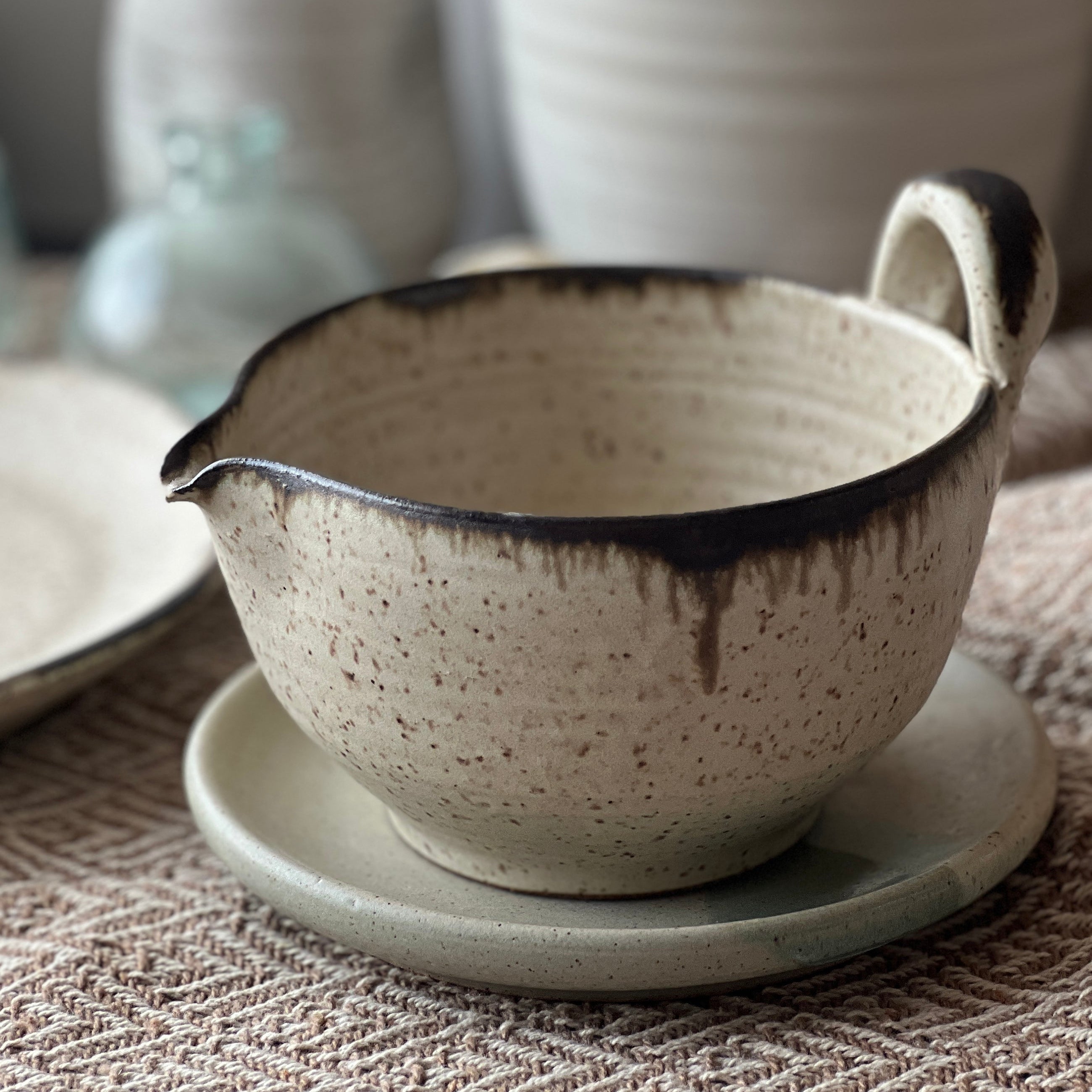 Ceramic bowl with handles on a textured surface, with other ceramic items in the background.