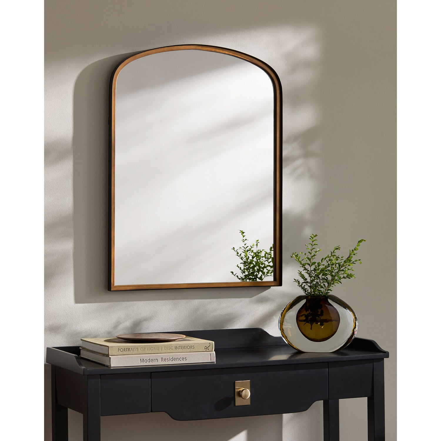 Wall mirror with wooden frame above a black console table with a vase and books.