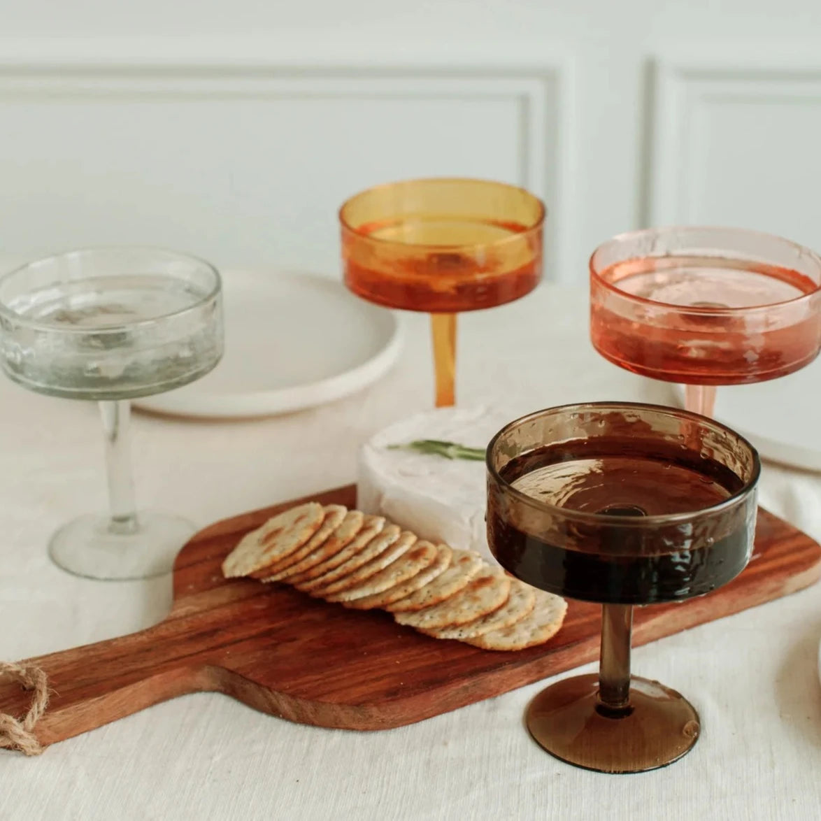 Cocktail glasses with different colored liquids on a wooden board with crackers.