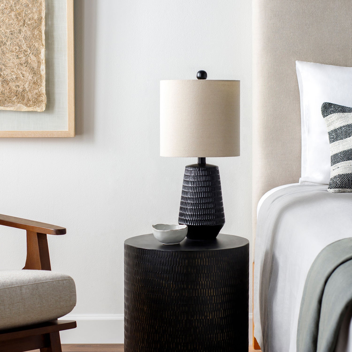 Black textured table lamp on a round black table next to a bed with striped pillow